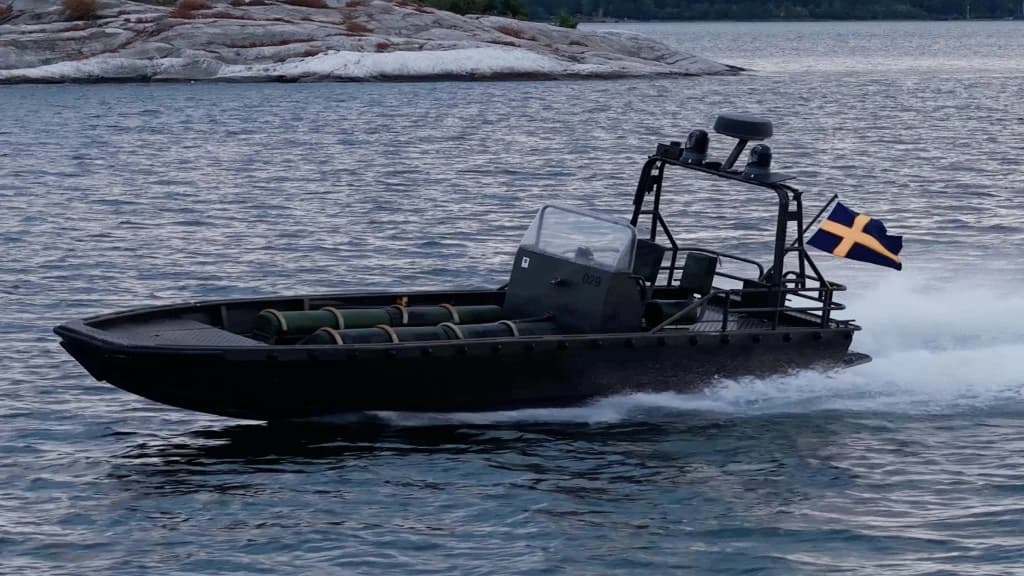 Nautrik vessel planing at speed past a rocky shoreline, Swedish flag flying
